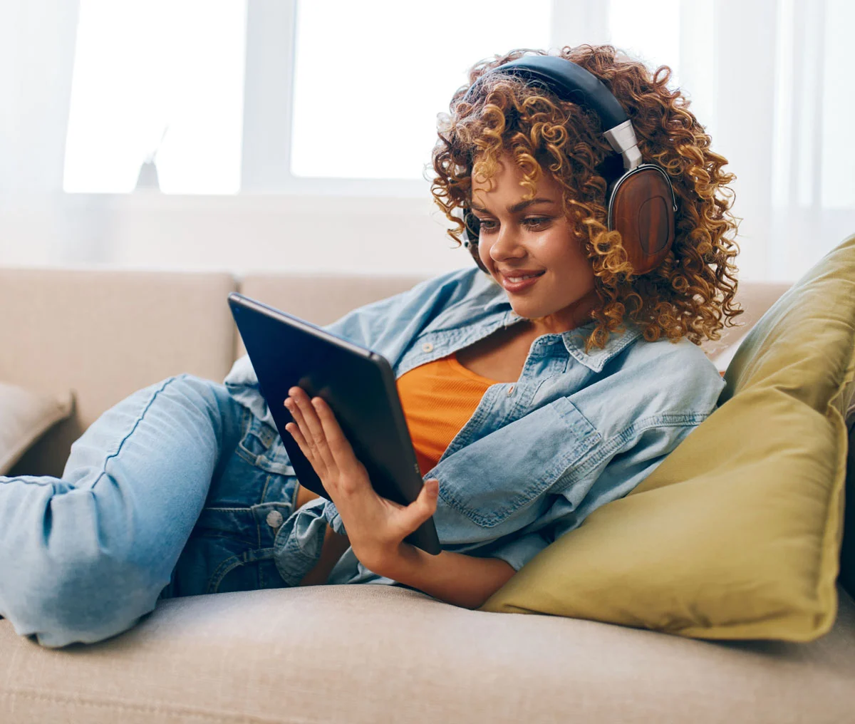 Mujer recostada en el sofá de su casa navegando en una tableta y con audífonos sobre su cabeza.